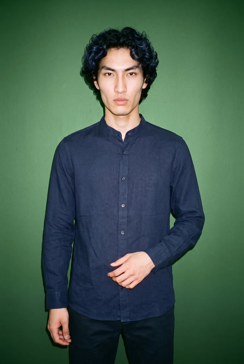 A 22-year-old Chinese male model wears a navy linen grandad shirt in a front-facing three-quarter-length view. He stands with his weight back and a natural facial expression against a hunter green studio-wall backdrop.