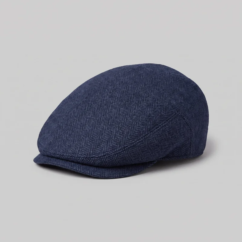 A complete navy flat cap is shown as a standalone product from a low three-quarter angle. The cap has a low rounded crown, curved panel seams, a short front brim, and an all-over fine herringbone pattern.