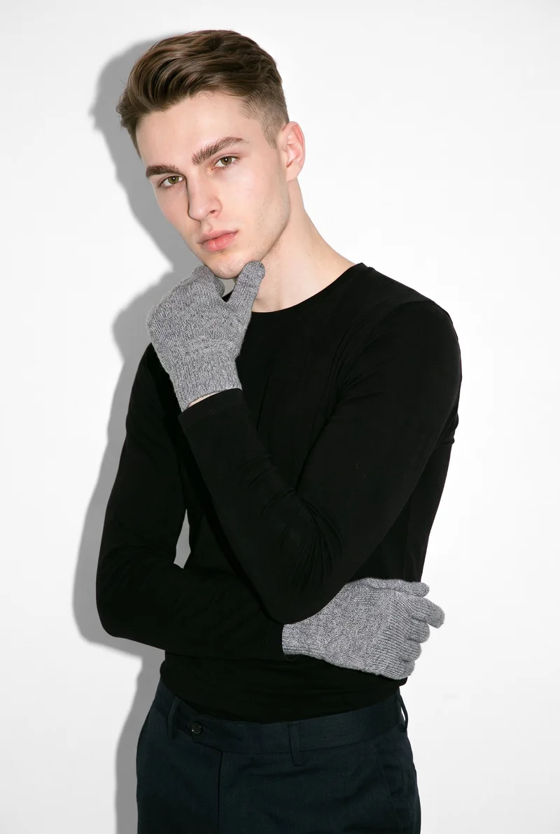A 21-year-old Caucasian man is shown from the upper body at an angled front view, wearing grey knit gloves with one wrist raised to his jaw. His expression is neutral, against a pure white studio backdrop.