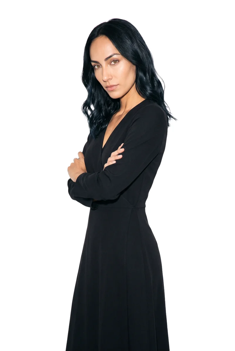 A 27-year-old Hispanic woman wears a black wrap dress in a three-quarter-length composition. She is shown from a front three-quarter angle in a self-hug pose with a curious facial expression, against a pure white backdrop.