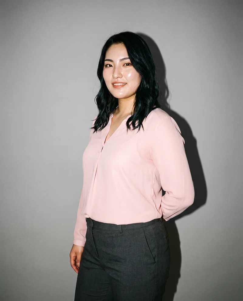 A 22-year-old Japanese woman wears a blush pink chiffon blouse in a three-quarter-length view. She is shown from a front three-quarter angle with one elbow positioned behind her back and a smile, against a pebble grey studio-wall backdrop.