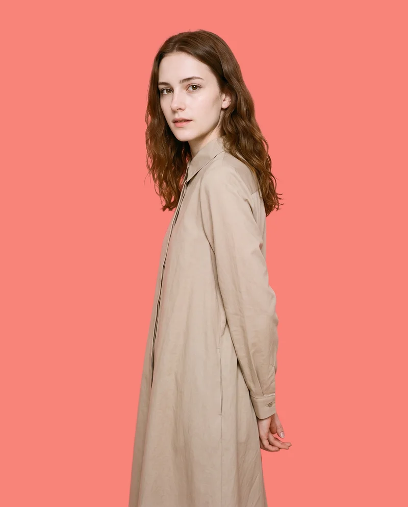 An 18-year-old Caucasian woman wears a beige modest shirt dress, shown in a three-quarter-length front three-quarter view with her hands behind her back and a natural facial expression. A flat coral pink backdrop frames the studio-cutout composition.