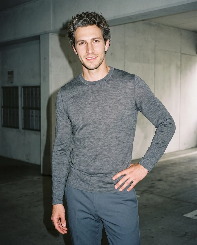 A 31-year-old Caucasian man wears a grey merino thermal top in a three-quarter-length frame. He is shown from a front three-quarter angle, smiling and shifting his weight back, with a concrete parking garage backdrop.