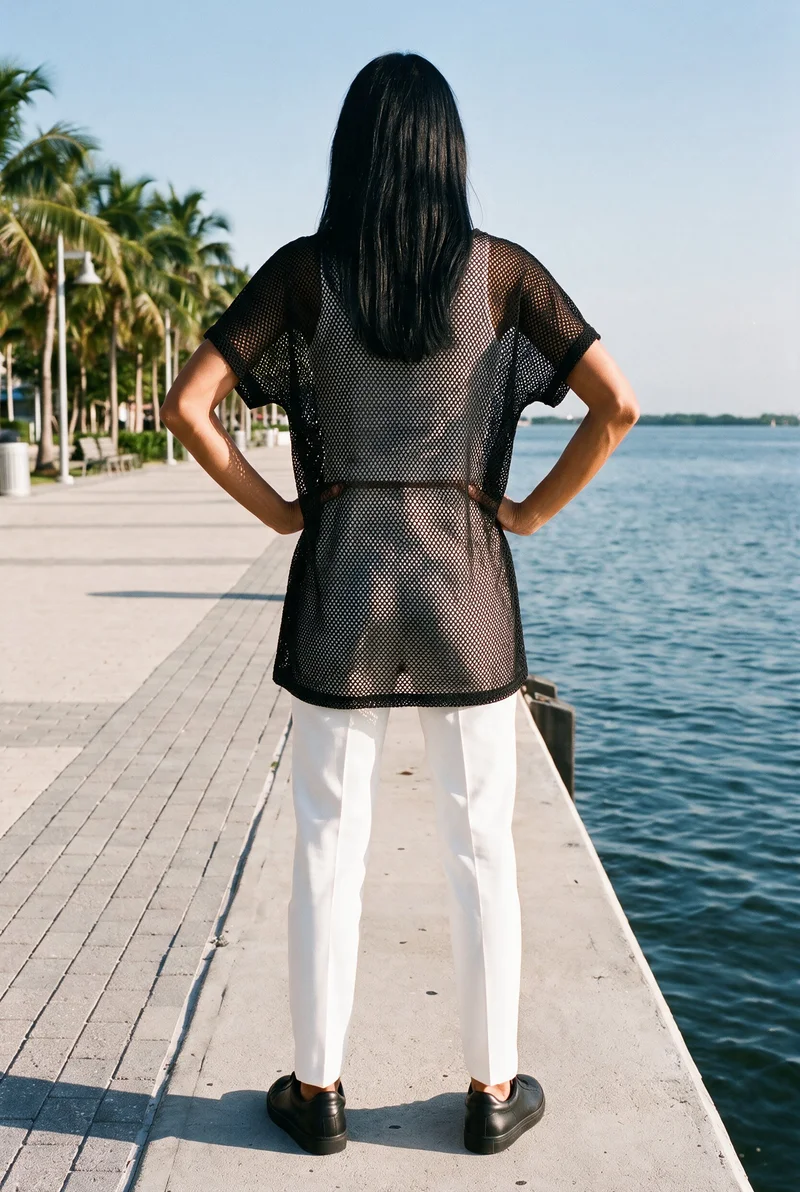 A 27-year-old Hispanic woman wears a women's black mesh cover-up in a full-body rear view, standing with her hands on her hips. Palm trees line the waterfront promenade backdrop.