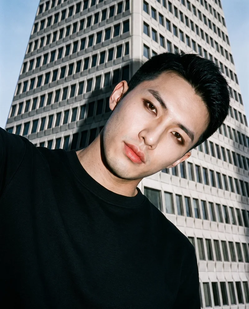 A 23-year-old Chinese man wears a black crewneck sweatshirt in a front-facing selfie with his head tilted and a neutral expression. A pyramid-shaped building appears as a low-angle backdrop.