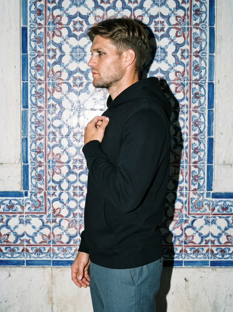 A 35-year-old Caucasian man is shown from the upper body in side view wearing a black pullover hoodie, with his fingers at his chest and a neutral expression. A Portuguese tile street serves as the backdrop.