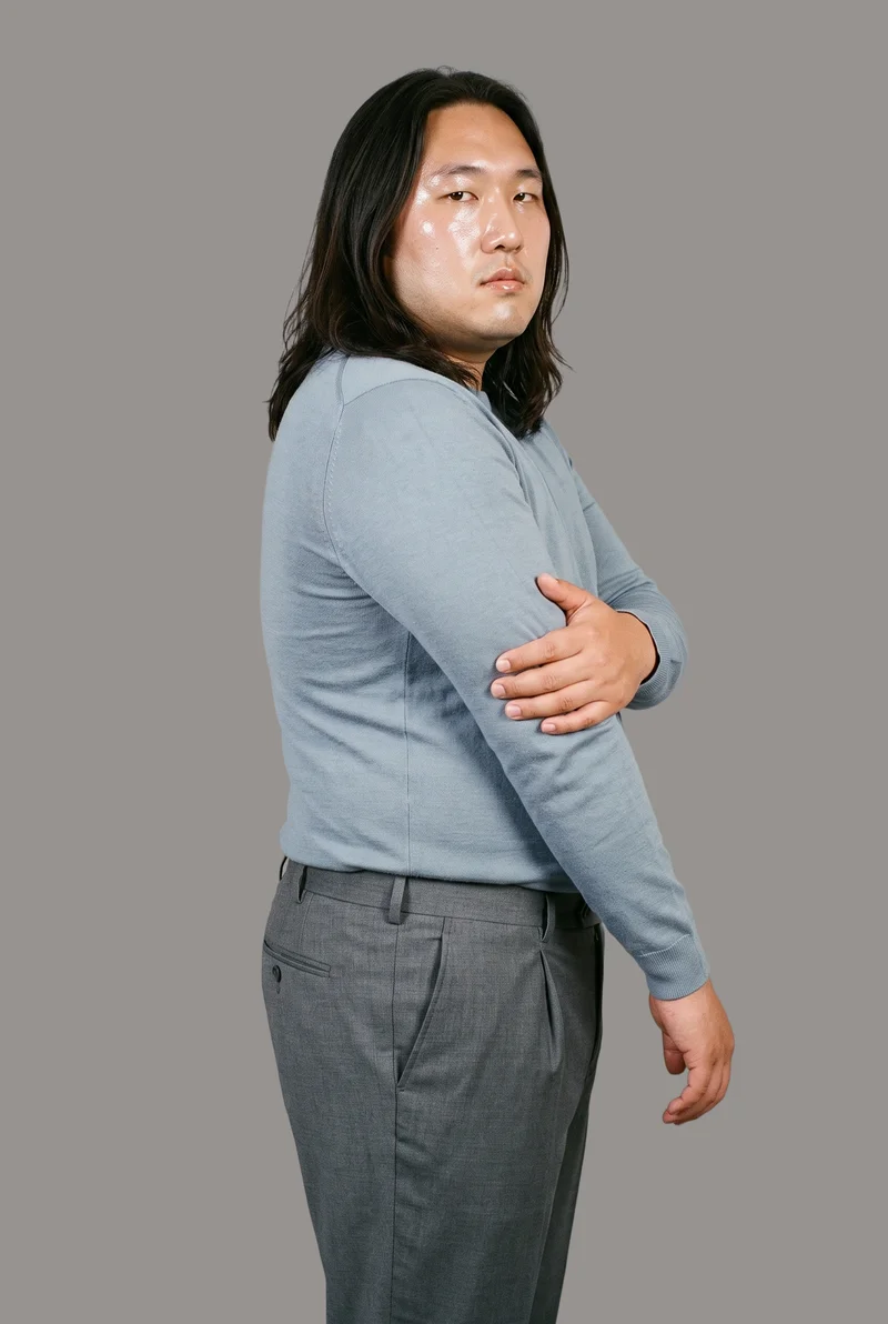 A 37-year-old Chinese man wears men's charcoal tailored trousers in a three-quarter-length frame, viewed from a front three-quarter angle. He is shown holding his arm with a confident expression against a flat studio-grey backdrop.