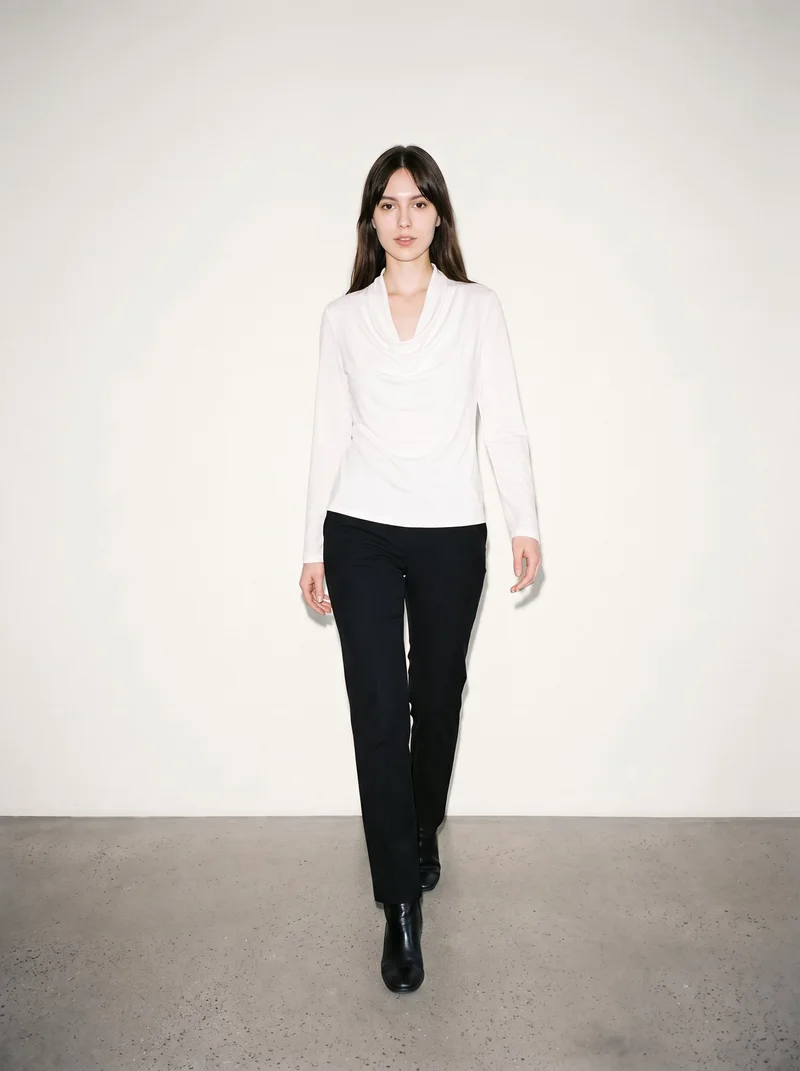 A 20-year-old Slavic woman is shown full body from the front while walking in a white draped cowl-neck top. She has a confident expression, with a soft ivory studio-wall backdrop.