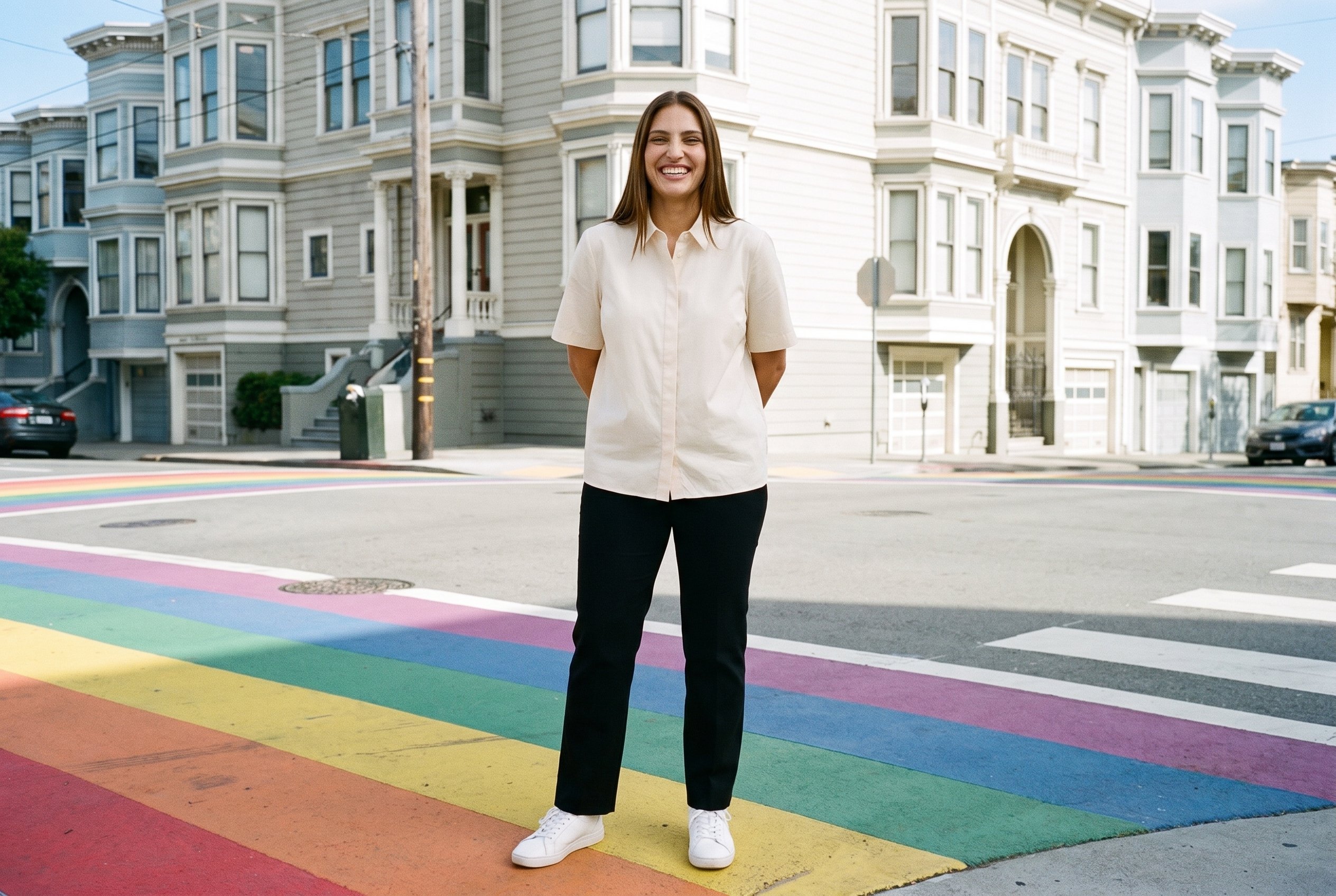A 31-year-old Hispanic woman wears a cream cotton blouse in a full-body front view, standing with her hands behind her back and grinning. The backdrop shows a rainbow crosswalk with Victorian houses.