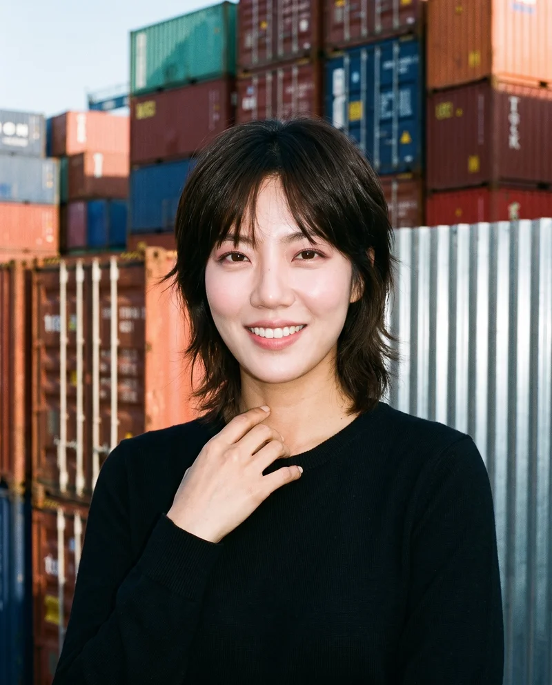 A 33-year-old Korean woman wears a black pullover jumper in a bust-length front view. She is smiling with one hand resting at her collarbone, against a backdrop of shipping containers.