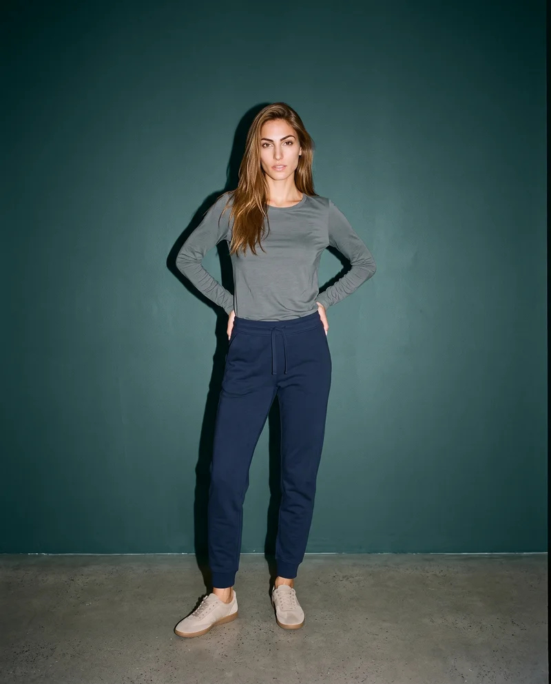 A 24-year-old Caucasian woman wears navy cotton joggers in a full-body front view with a wide stance and confident expression. A dark slate studio-wall backdrop frames the composition.