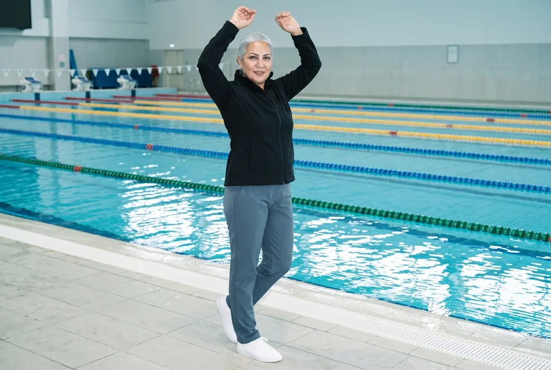 A 46-year-old Turkish woman wears a black softshell jacket in an environmental front three-quarter view, with her arms raised overhead and a natural expression. An Olympic swimming pool serves as the backdrop.