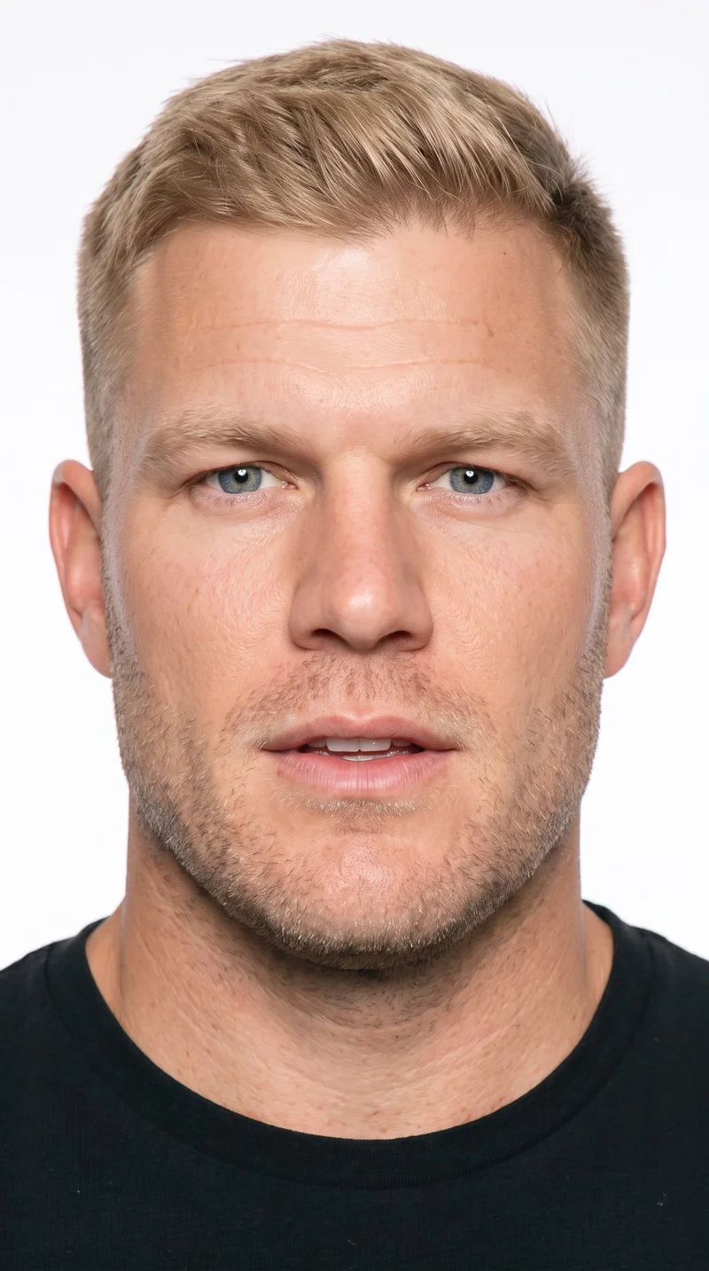 A 36-year-old Caucasian man with lightly tanned fair skin and a peach undertone. His rugged face has weathered, characterful features and a heavy jaw, with thin eyebrows, blue eyes, and thin lips. He wears blonde hair in a two-block style with short sides and back and a longer, fuller top, along with fine, closely trimmed stubble over the beard area.