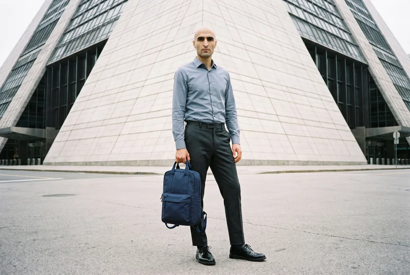 A 37-year-old Slavic man is shown full body from the front with a neutral expression, displaying a men's navy canvas backpack as it dangles. A pyramid building appears as a low-angle backdrop.