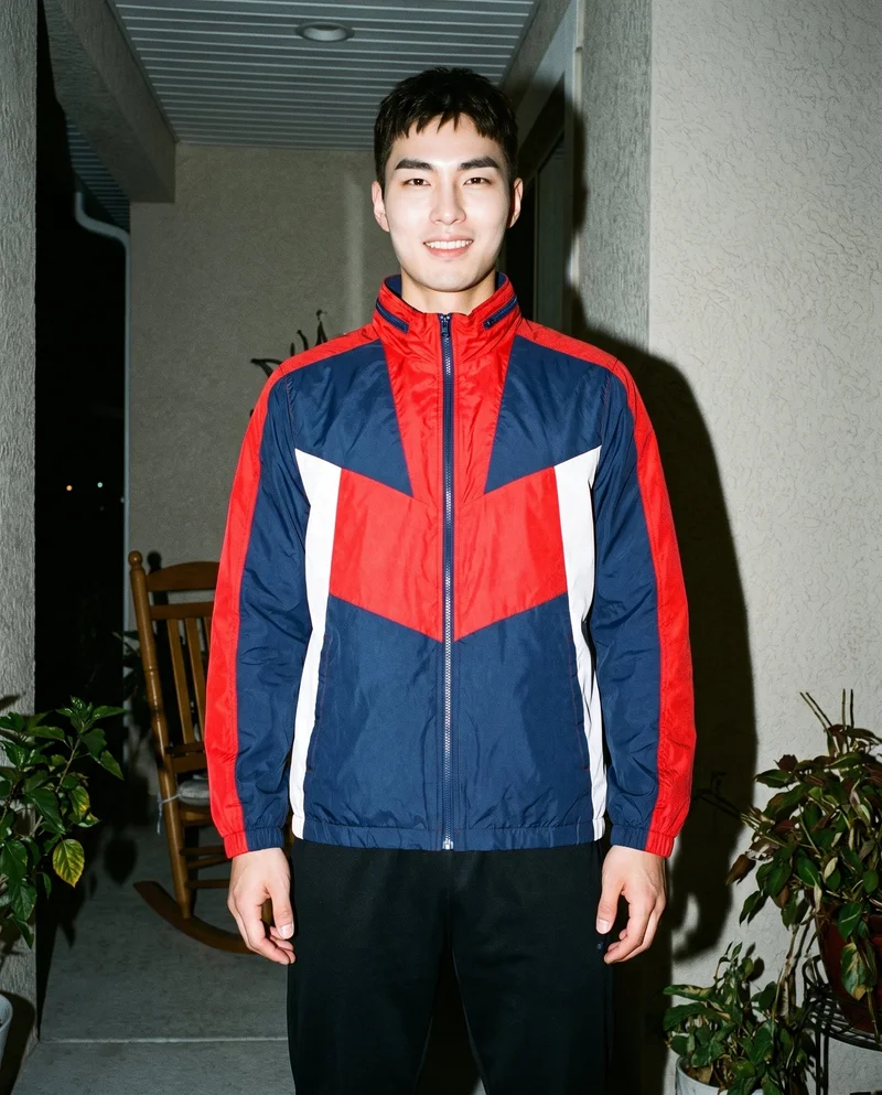 A 26-year-old Korean man stands straight in a front-facing, three-quarter-length view wearing a men's red colour-block windbreaker. His natural expression is visible against a front porch backdrop.