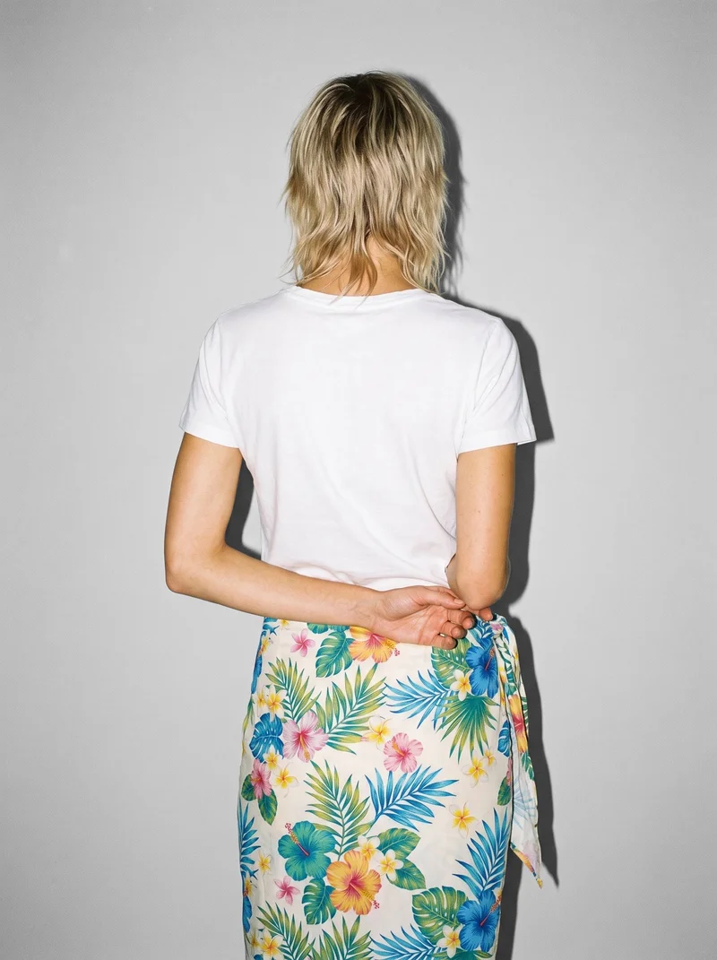 A 22-year-old Caucasian woman models a women's floral print sarong in a three-quarter-length rear view, standing with her hands behind her back. A Fog Grey studio-wall backdrop frames the composition.