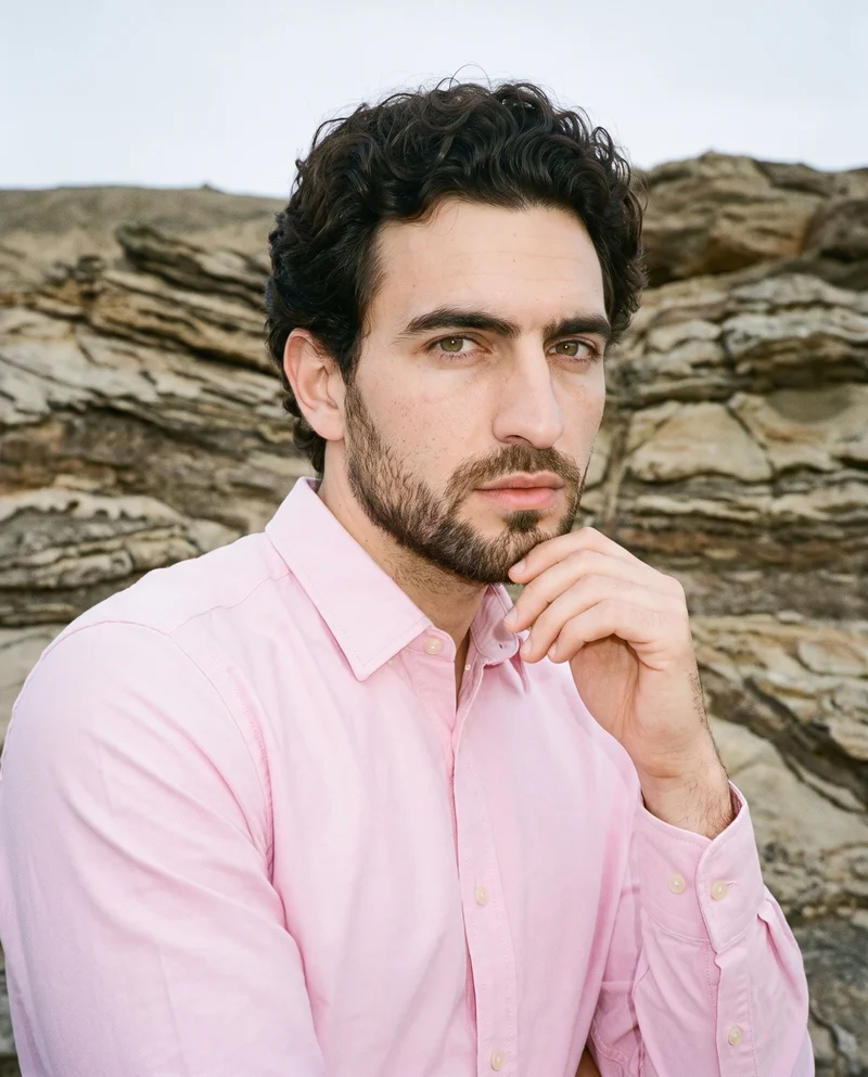 A 33-year-old Caucasian man wears a pink Oxford shirt in a bust-length front three-quarter view, with one hand resting at his jaw and a confident expression. A Badlands backdrop is visible behind him.