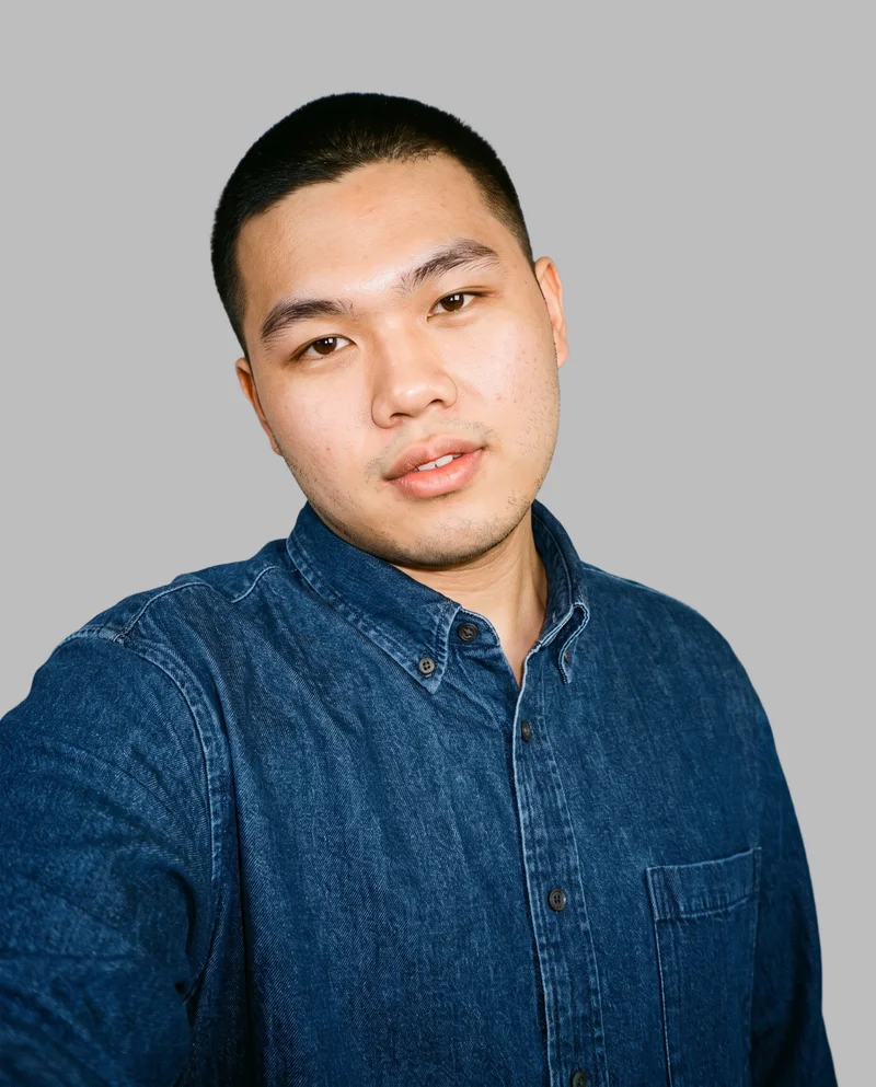 A 20-year-old Southeast Asian man wears an oversized denim shirt in a front-view selfie-style composition. His head is tilted and his expression is neutral, with a dove grey flat-color backdrop.