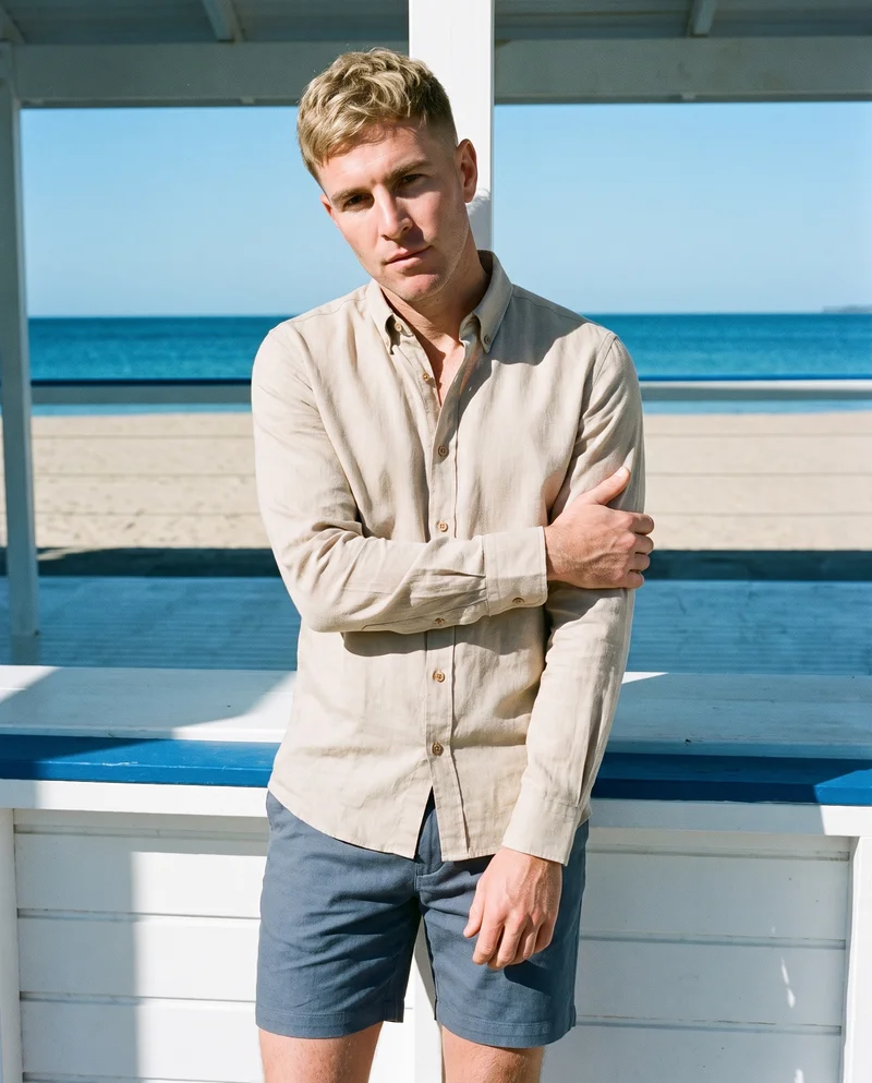 A 40-year-old Caucasian man wears a beige linen shirt in a 3/4-length front 3/4 view. He holds one arm in a natural pose, with a Greek island beach club backdrop.