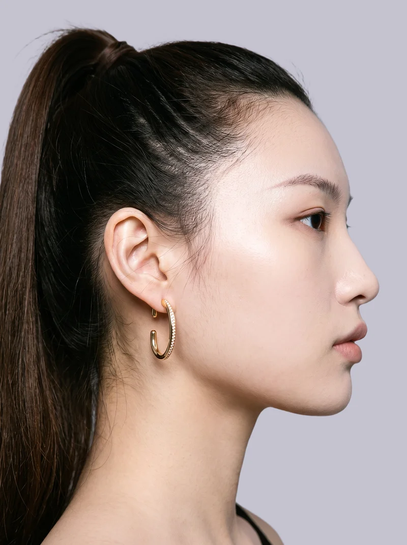 A gold ear cuff is shown on an ear in side view, with the ear positioned forward. The lavender-grey flat-color backdrop keeps the focus on the accessory.