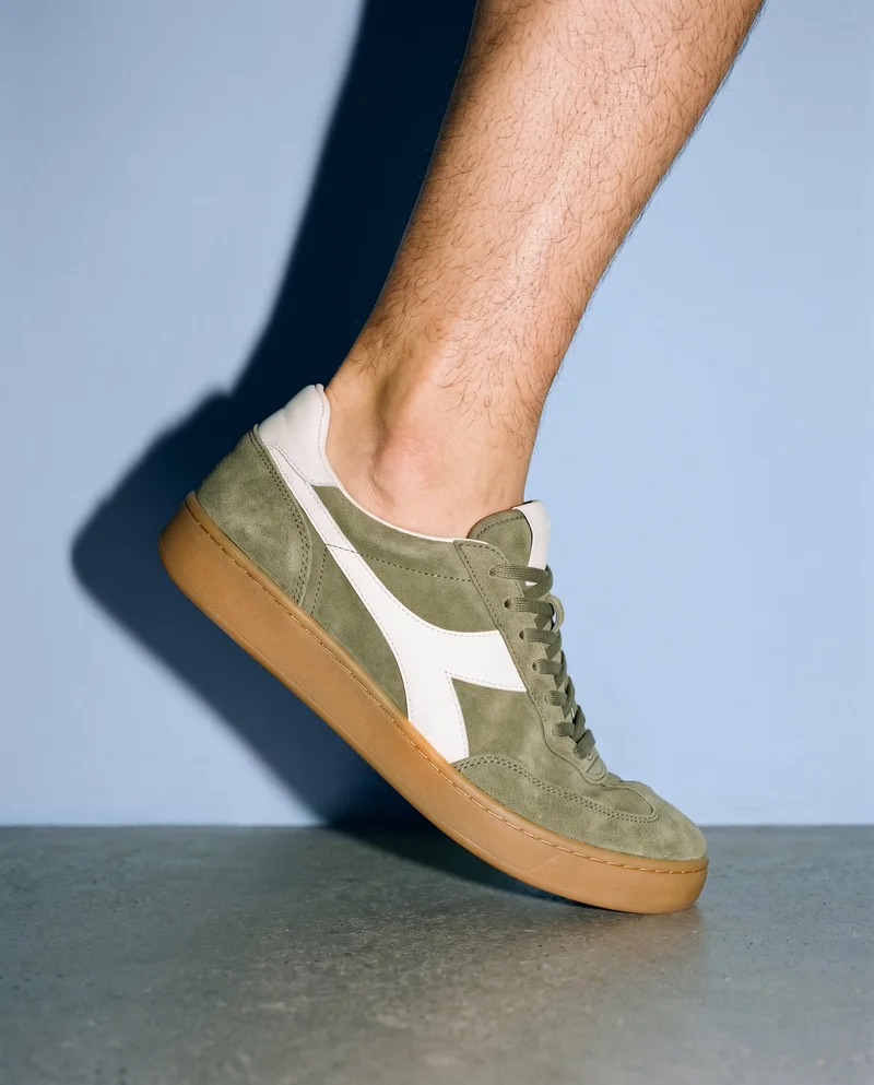 Men's retro suede sneakers shown from the side as they are worn on tiptoes, framed from the ankles down against a powder-blue studio-wall backdrop.