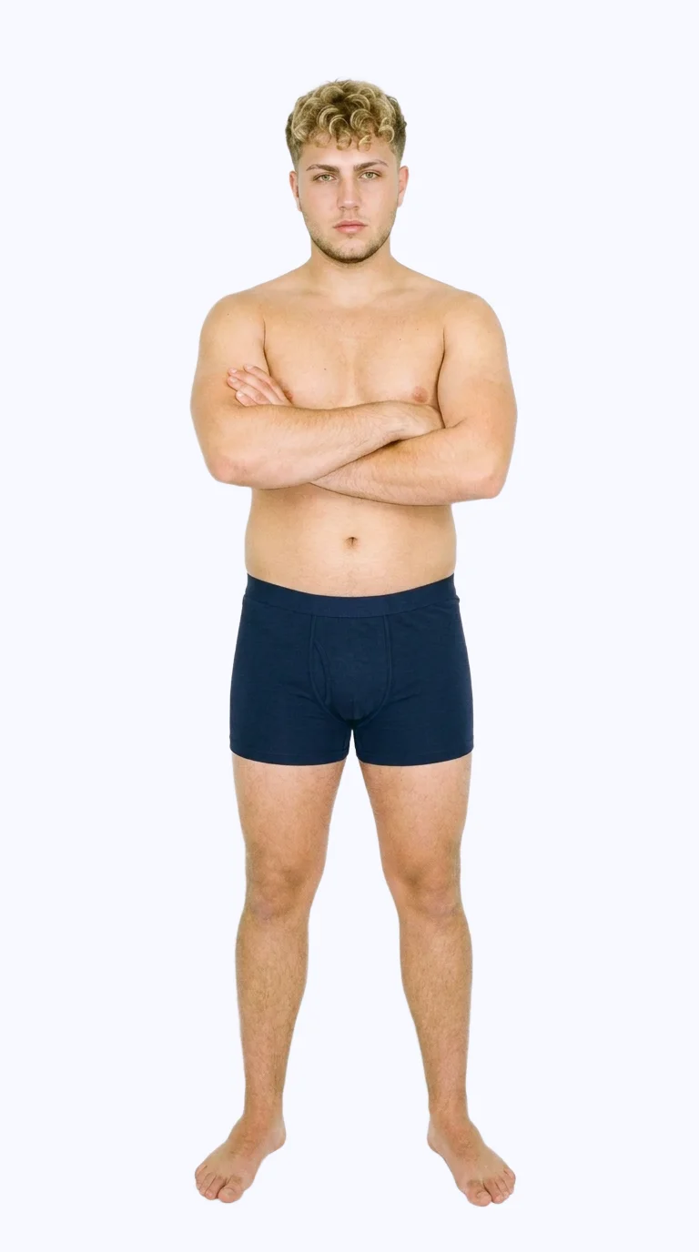 An 18-year-old Caucasian male model wears navy cotton trunks in a full-body front view, standing with his arms crossed and a neutral facial expression. A flat porcelain-colored backdrop frames the studio cutout.