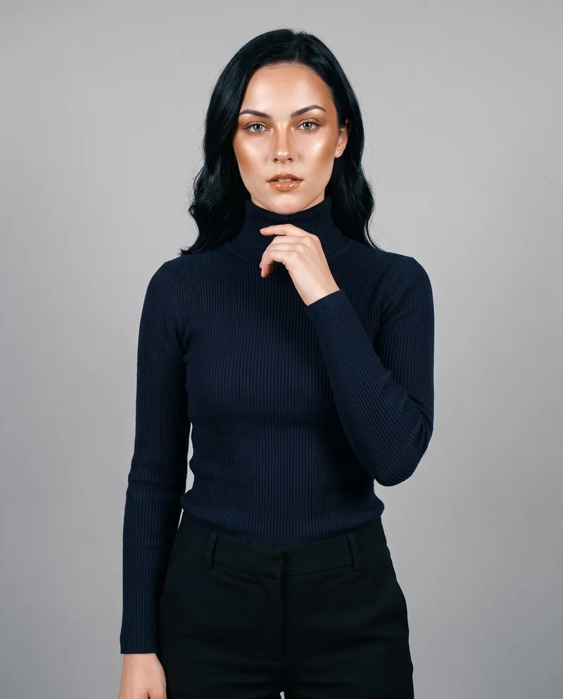A 19-year-old Caucasian woman is shown from the upper body in a front view, wearing a navy ribbed turtleneck with one hand at her collarbone and a neutral expression. A Silver Mist studio-wall backdrop fills the background.