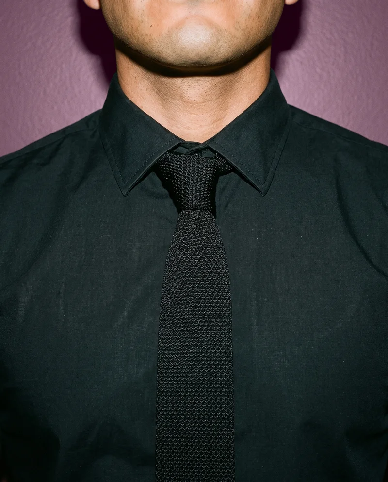 A black knitted tie is shown from the front at neck level, with the neck area framed against an eggplant studio-wall backdrop.