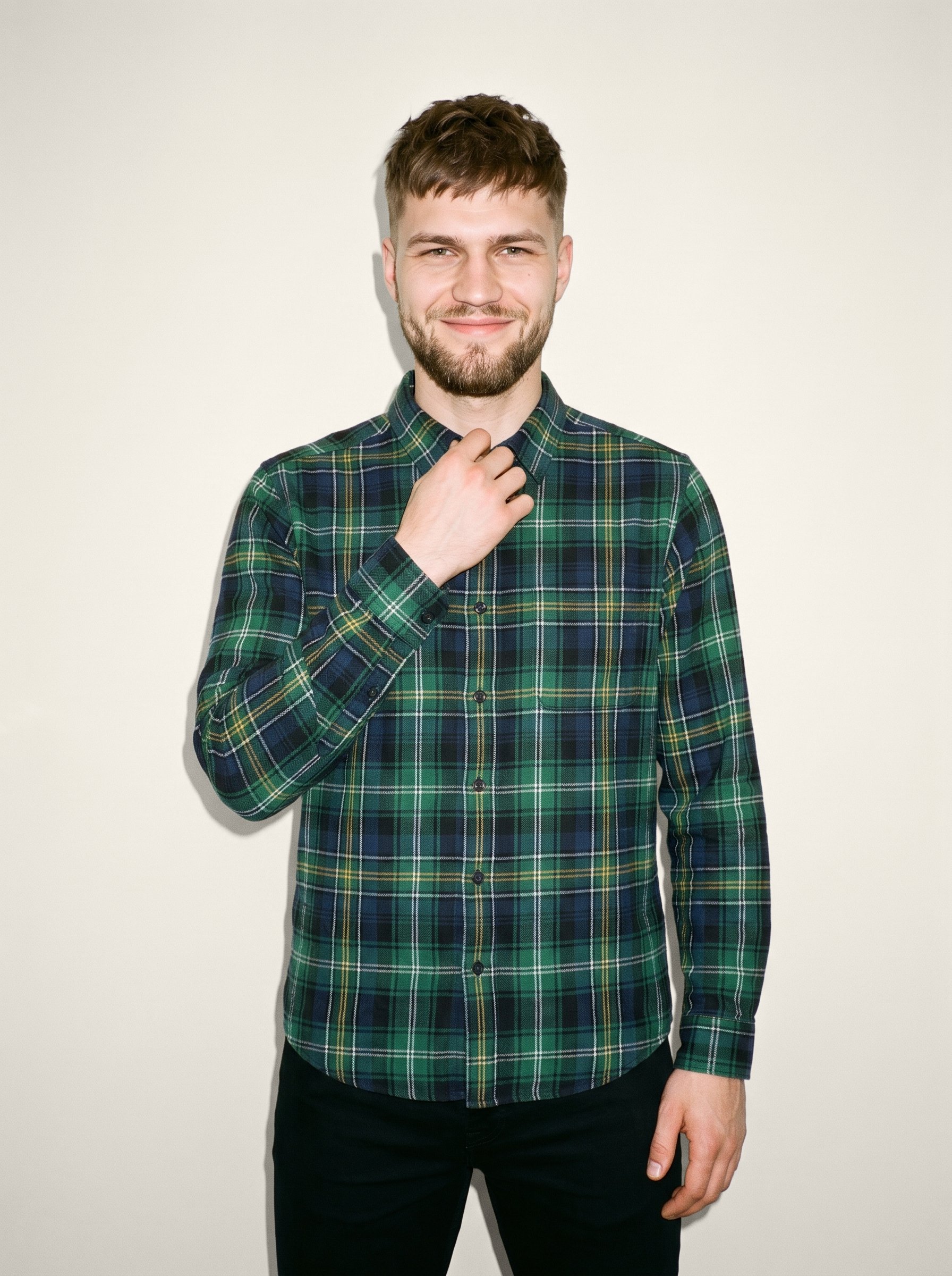 A 22-year-old Slavic man is shown from the front in an upper-body view, wearing a green plaid flannel shirt with one hand at his collarbone. His natural expression is visible against an Oyster Pearl studio-wall backdrop.