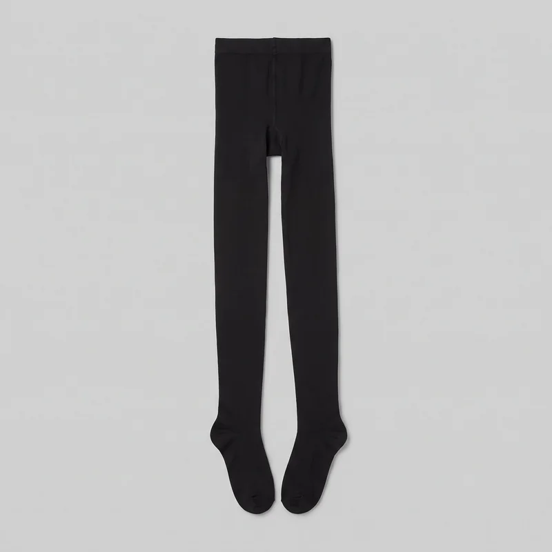Black opaque footed tights are laid out flat in a complete product view. The full-length design includes a wide waistband, a vertical center seam through the crotch, a shaped gusset, subtle leg seams, integrated foot sections, and rounded toe ends.