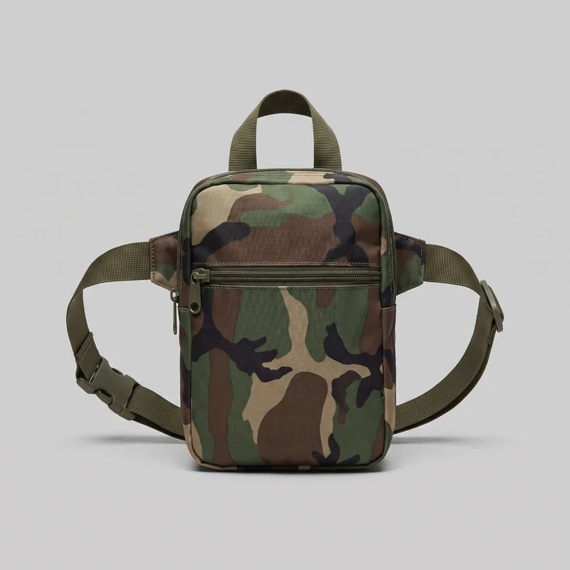 A woodland camouflage utility bum bag is shown in a mostly complete front view. The vertical rectangular pouch has a squared base, rounded upper corners, side gusset panels, and raised piping around the front edges. A horizontal zip pocket with a visible pull spans the upper front, while a top zip track, short webbing grab handle, adjustable wide webbing strap, slider hardware, and side-release buckle are visible.