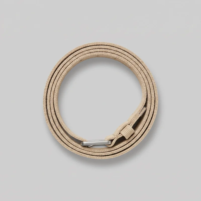 A beige canvas belt is shown coiled into a round shape in a top-down standalone view. The flat, flexible strap has a fine ribbed woven texture and lengthwise stitched or bound-looking edges, with a rectangular metal buckle or slide bar, matching keeper loop, and narrow metal tab visible near the coil.