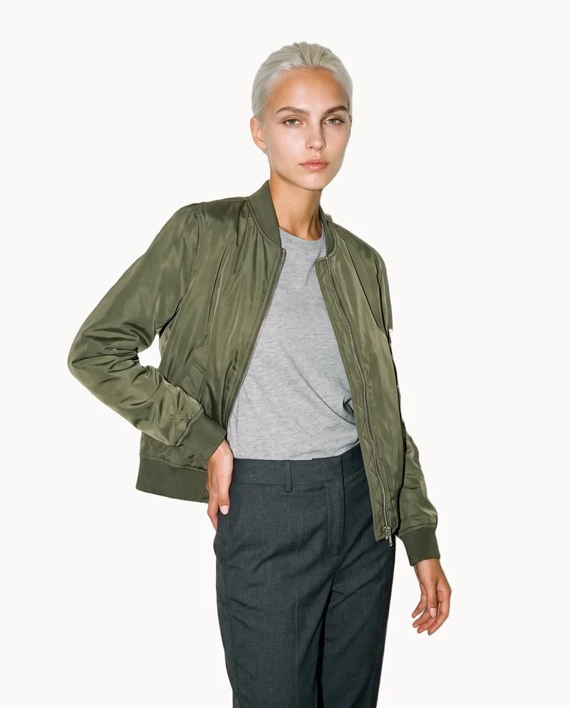 A 22-year-old Caucasian woman wears an olive MA-1 bomber jacket, shown from a front three-quarter angle in a three-quarter-length frame. She stands with her weight positioned back and a soft facial expression against a Chantilly Lace flat-color backdrop.
