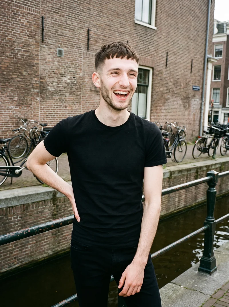 A 20-year-old Caucasian man wears a black T-shirt in an upper-body front view, laughing with one hand on his hip. A canal bridge with bicycles forms the backdrop.