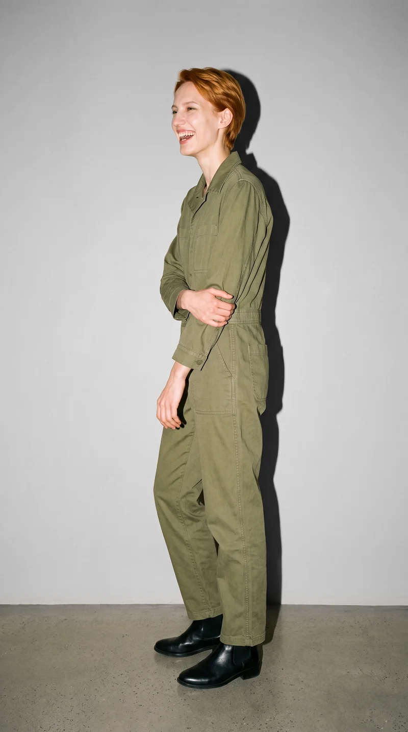 A 21-year-old Caucasian woman wears an olive workwear boilersuit in a full-body side view, standing with her arms low and laughing. A dove grey studio-wall backdrop fills the background.