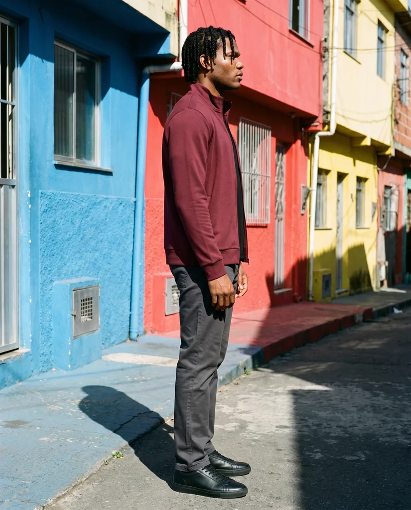 A 26-year-old African man wears a burgundy tracksuit top in a full-body side view, standing with his hands behind his back and a natural expression. A Brazilian favela street serves as the backdrop.