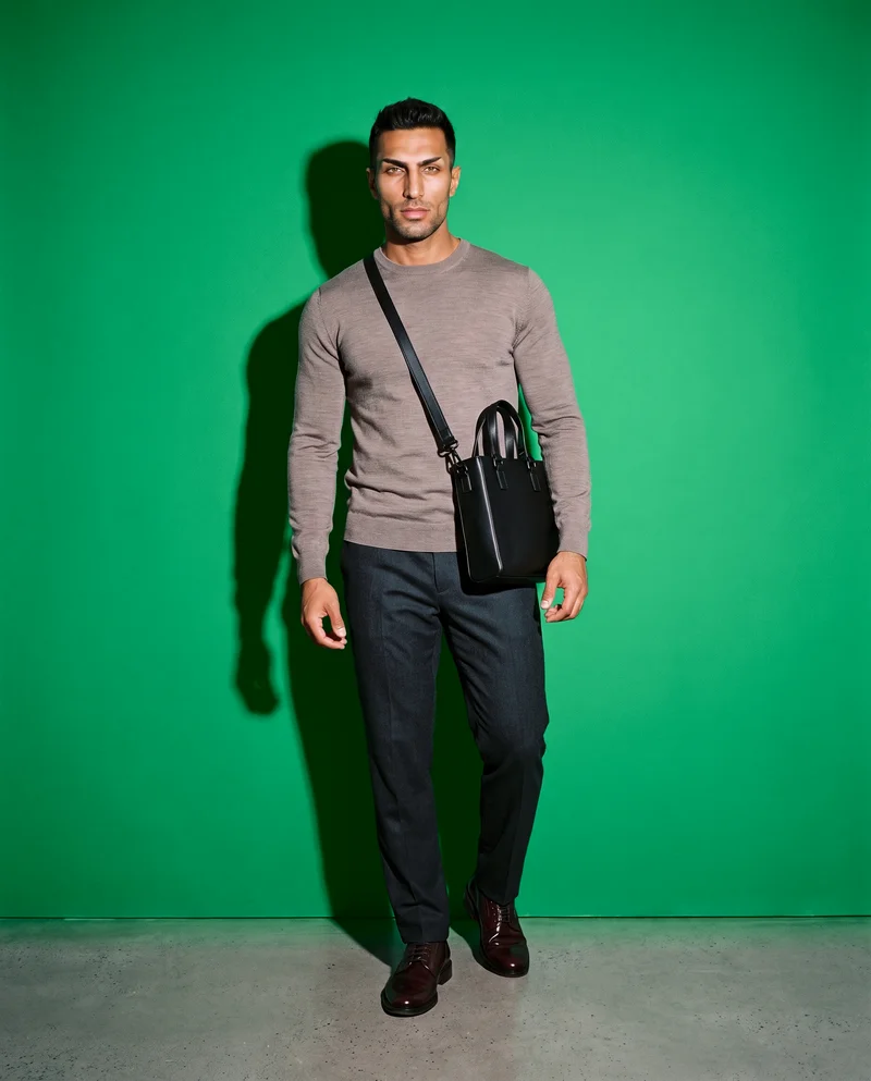 A 31-year-old Arab man stands in a relaxed pose, shown full body from the front, carrying a men's black leather crossbody bag. He has a confident expression, with a jade green studio-wall backdrop.