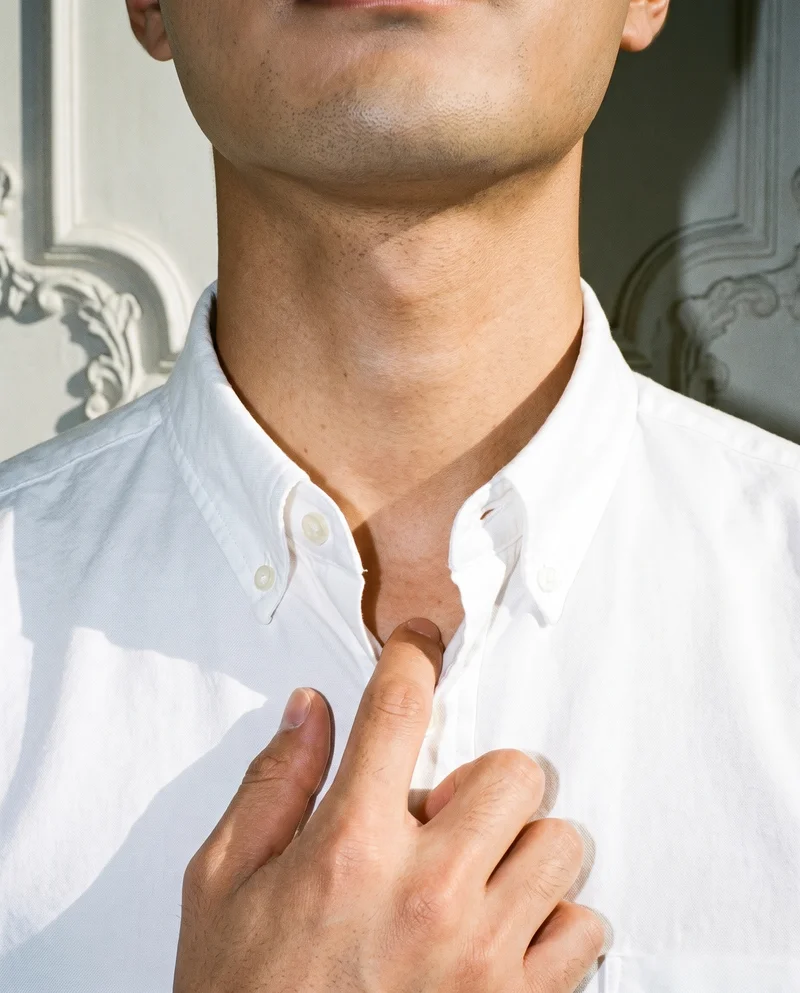 A man wears a white Oxford shirt in a front-view crop framed at the neck, with one finger positioned at his throat. A mansion foyer serves as the backdrop.