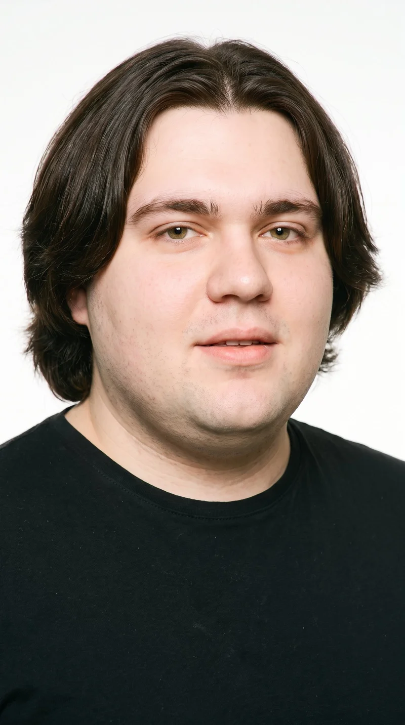 This 23-year-old Caucasian man has fair skin with a peach undertone and a round face with soft, full contours and few angles. His eyebrows are medium in thickness, and his hazel eyes contain green-gold flecks. He has full lips, dark-brown hair divided at the centre and falling evenly to both sides, and no facial hair, leaving his face smooth.