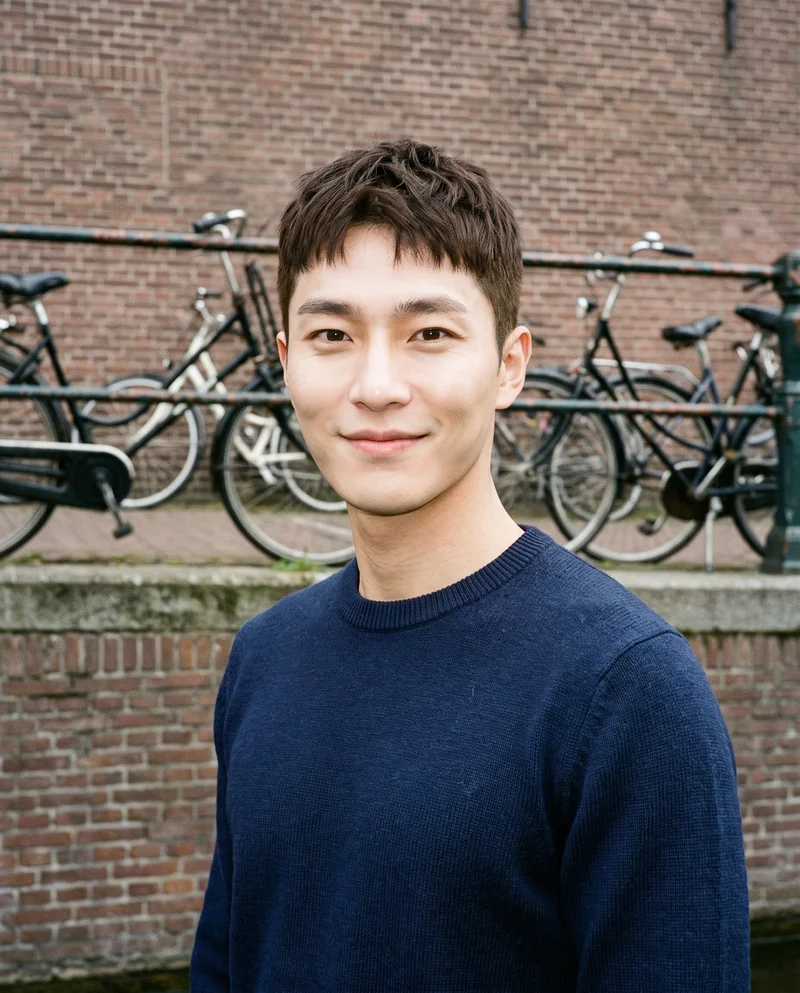 A 30-year-old Korean man wears a navy wool pullover jumper in a bust-length view. He is shown from a front three-quarter angle with his head level and a smile, against a Canal Bridge Bikes backdrop.