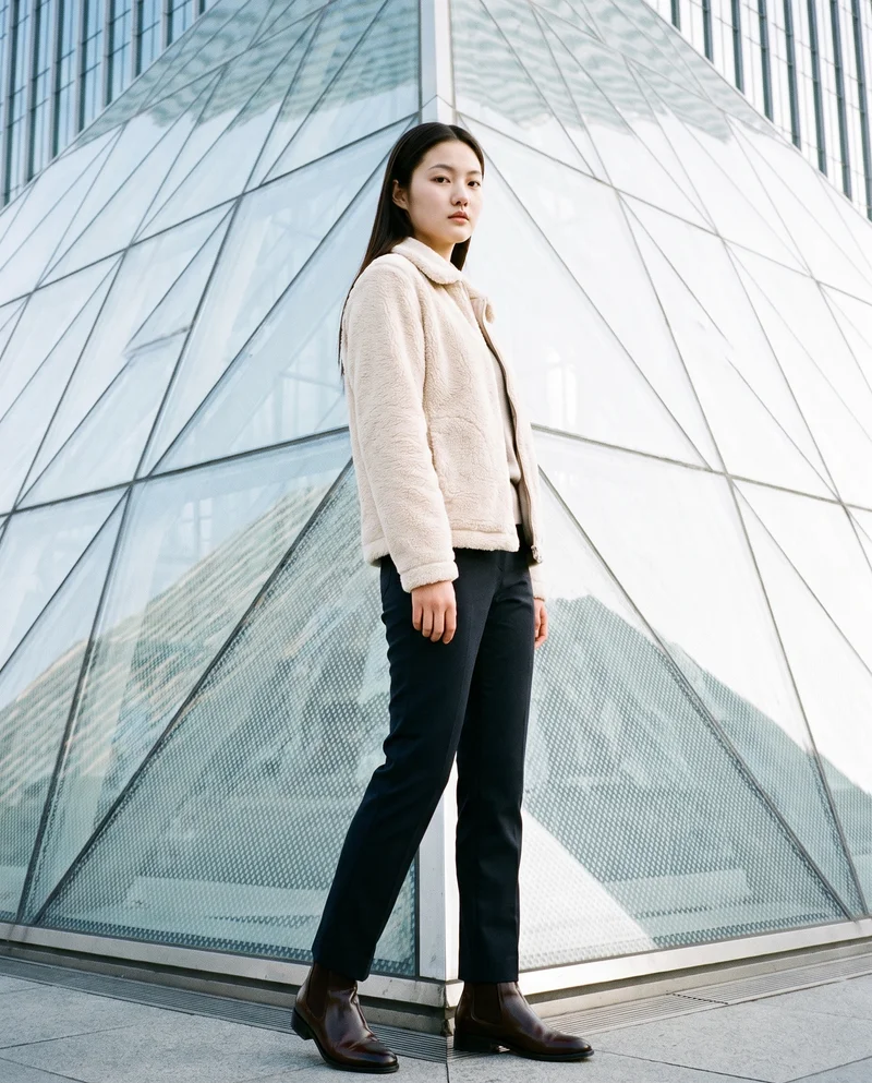An 18-year-old Chinese woman wears a cream sherpa fleece jacket in a full-body view from a front three-quarter angle. She stands with her weight back and a neutral facial expression, with a pyramid building shown as the backdrop.