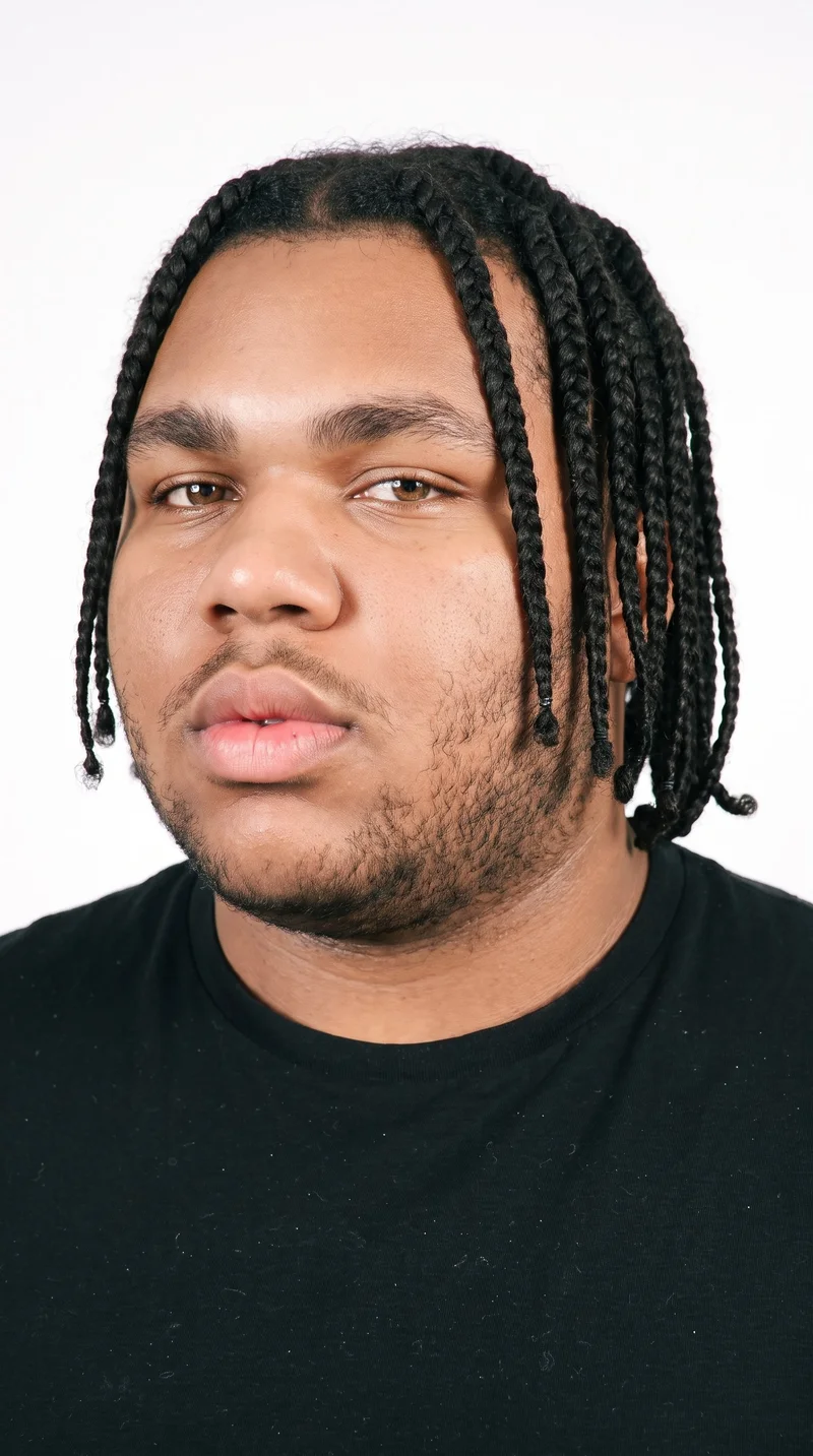 A 20-year-old African man with medium-brown skin and a cool undertone has a softly refined, even face shape. His thick eyebrows frame hazel eyes with green-gold flecks, and his lips are medium in fullness. His deep black hair is styled in long individual box braids formed from square sections. Short, even three-day beard growth adds a clearly visible, closely groomed texture along the lower face.