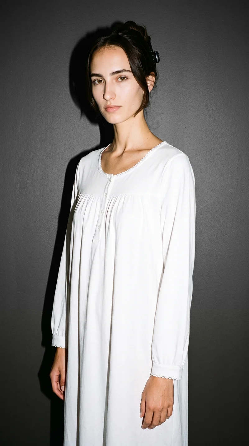 A 20-year-old Caucasian woman models a white cotton nightdress in a three-quarter-length front three-quarter view. She stands with her weight back and a serious expression against a charcoal slate studio-wall backdrop.