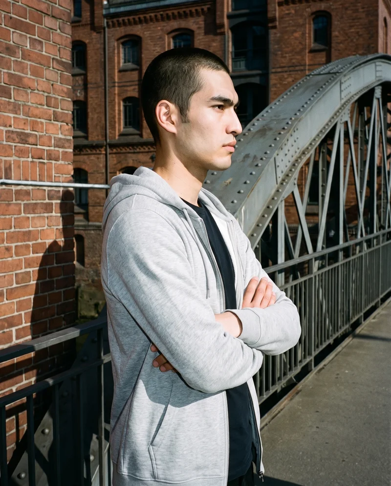 A 23-year-old Japanese man wears a grey tracksuit top in an upper-body side view with his arms crossed and a soft facial expression. A bridge-framed urban view forms the backdrop.
