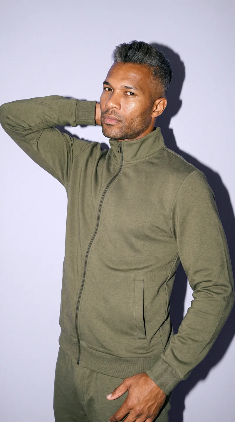 A 36-year-old African man wears an olive tracksuit in an upper-body view. He is shown from a front three-quarter angle with one hand behind his neck and a neutral facial expression, against a soft lavender studio-wall backdrop.