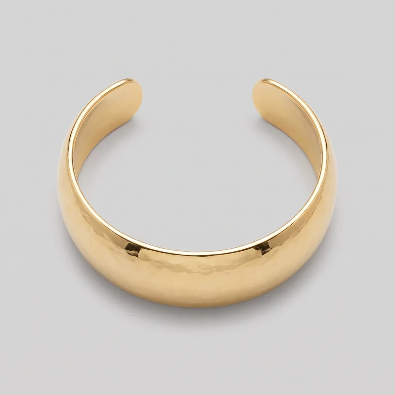 A complete wide gold open cuff bracelet is shown alone from an angled top view. Its rigid crescent-shaped band has a broad domed profile, a rounded open gap at the top, blunt tapered terminals, subtle irregular hammered-like undulations, and a narrow raised rounded rim along the inner circumference near both ends. No clasp, stones, charms, logos, prints, or other embellishments are visible.