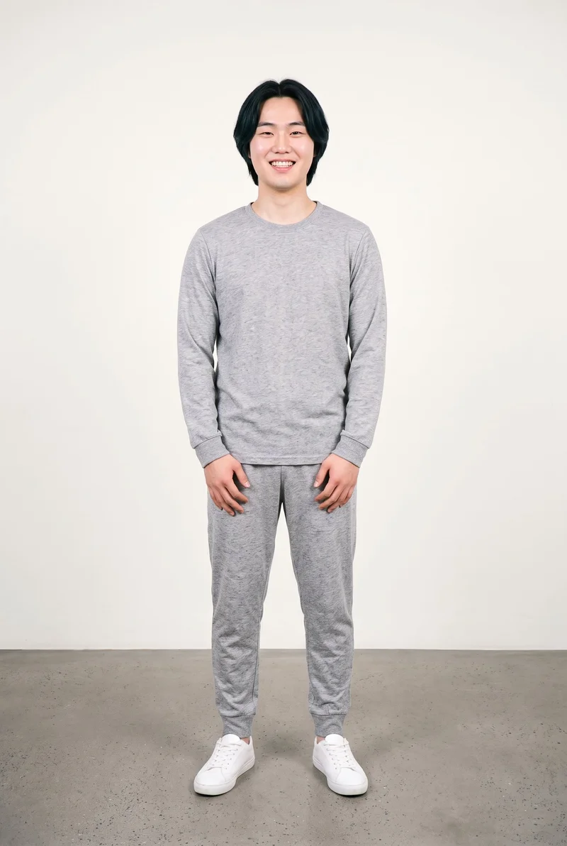 A 25-year-old Korean man models a men's grey marl pyjama set in a full-body front view, standing with his hands in front and grinning. A soft ivory studio-wall backdrop frames the outfit.