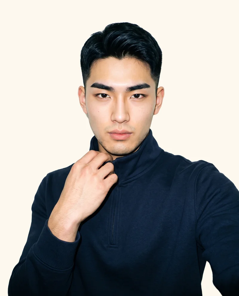 A 19-year-old Japanese man wears a navy quarter-zip in a front-view, over-the-shoulder selfie. His fierce expression is framed against a creamy white flat-color backdrop.