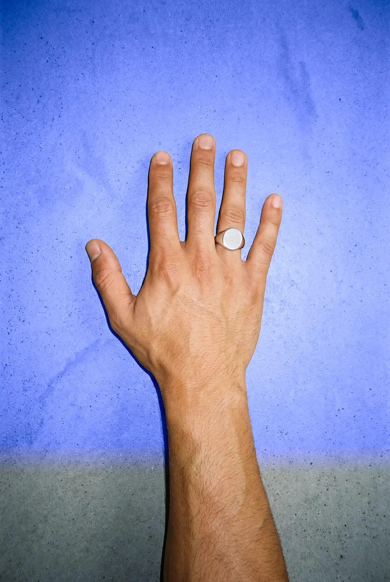 A silver signet ring worn on a hand shown flat from a top view, with the wrist area included in the frame. The image uses a cobalt studio backdrop.