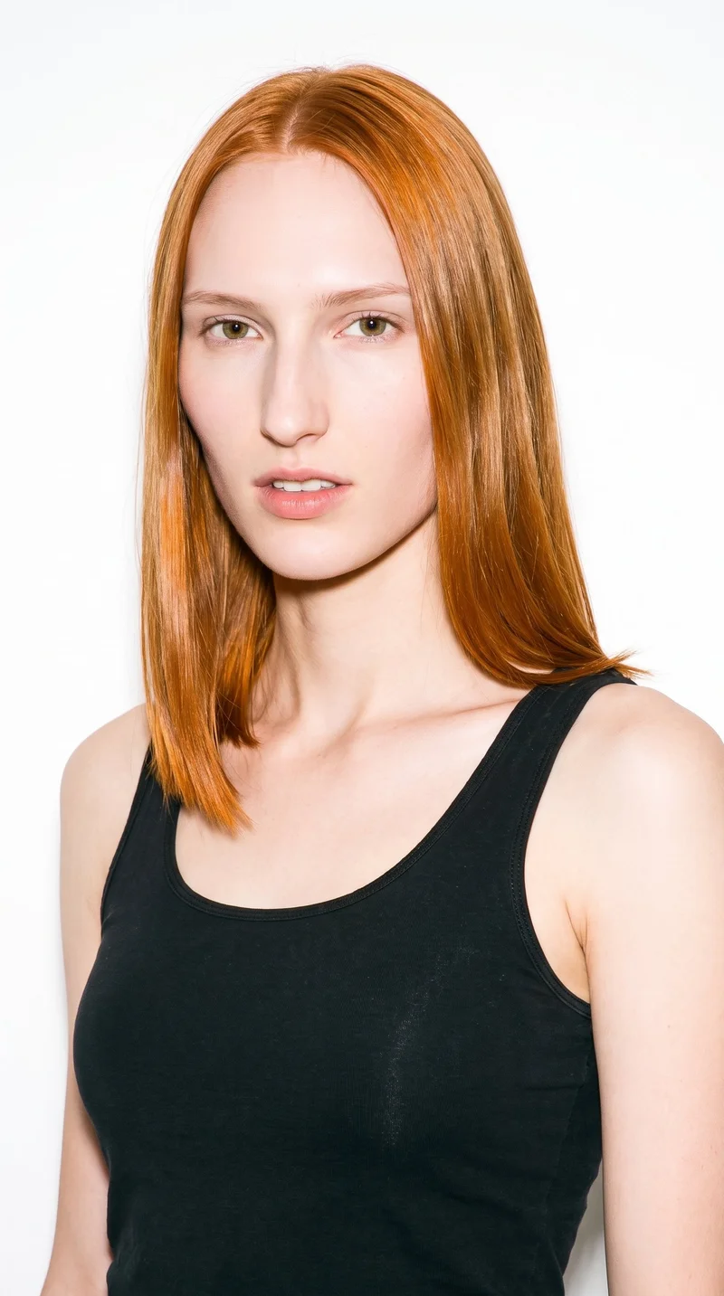 This 19-year-old Caucasian woman has fair skin with a warm golden undertone. Her face is long, with more length than width, a tall forehead, and elongated cheeks. She has medium-thickness eyebrows, green eyes, and medium-sized lips. Her copper-red hair is straight, smooth, and divided evenly at the centre.